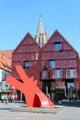 Abstract sculpture & shop with red grating facade before Cathedral spire in Market Area. Ulm, Germany.
