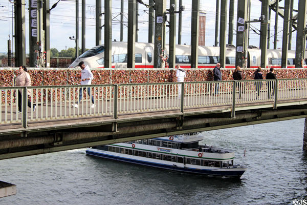 Love locks attached to Hohenzollern Bridge with passenger train crossing and tour boat passing underneath. Köln, Germany.