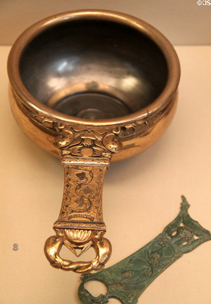 Roman era copper alloy skillet features dolphins & sea creatures (2ndC CE) by Boduogenus from Isle of Ely, Cambridgeshire beside Roman bronze pan handle at British Museum. London, United Kingdom.